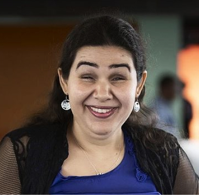 Luciana esta sorrindo. Ela esta com o cabelo preso, com um batom vermelho e brinco de prata.